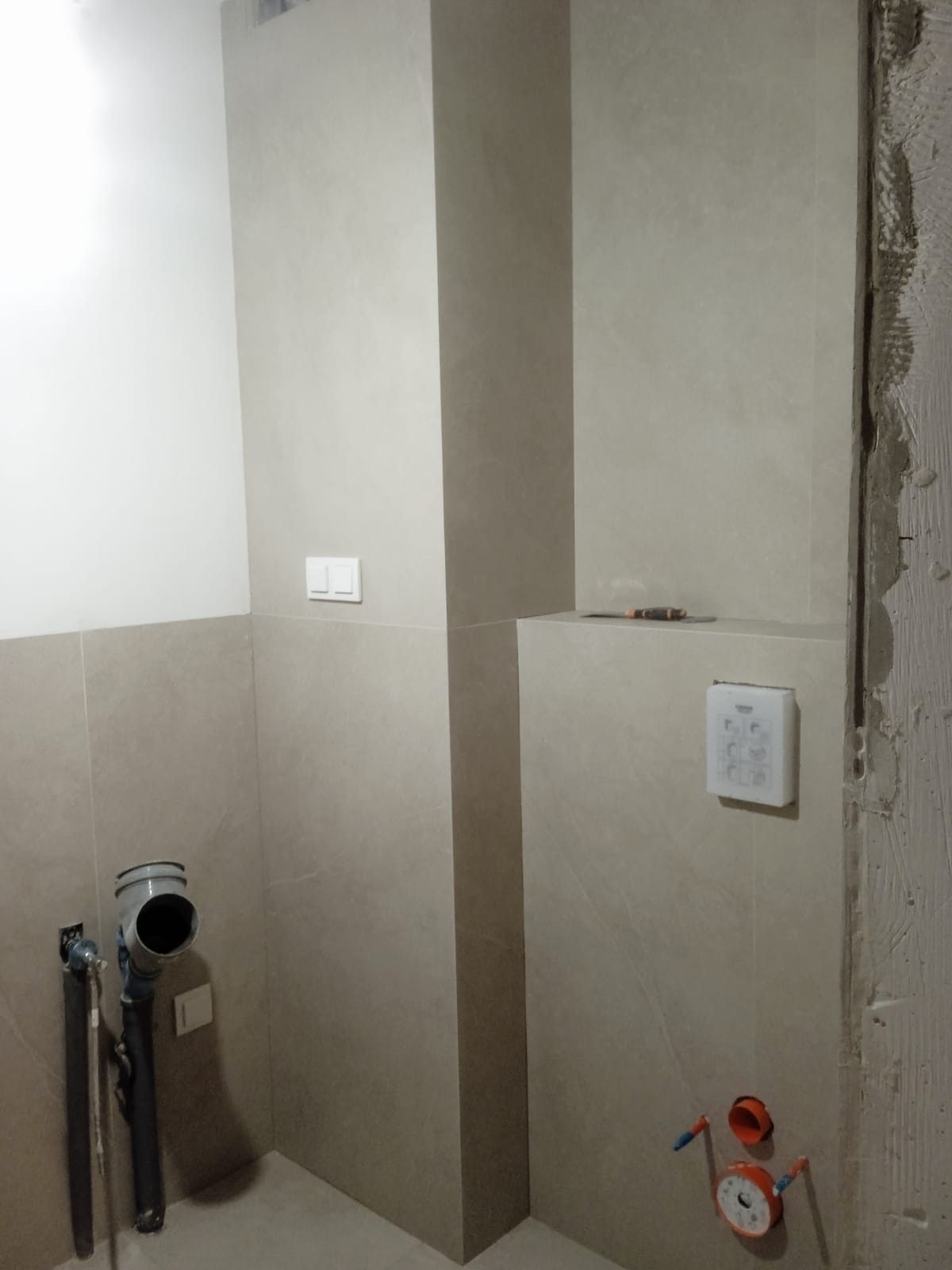 Ściana łazienki w trakcie remontu z beżowymi płytkami, widoczne rury kanalizacyjne, przycisk spłuczki oraz narzędzie na półce.