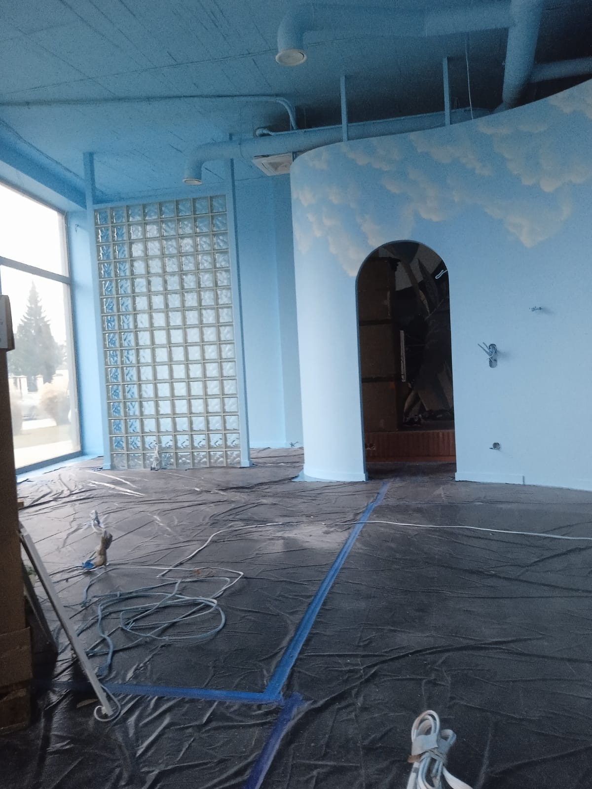 Wnętrze lokalu w trakcie prac malarskich, zabezpieczona podłoga folią i taśmą malarską, świeżo pomalowane na niebiesko ściany i sufit, dekoracja malarska chmur na ścianie, ściana z luksferów.