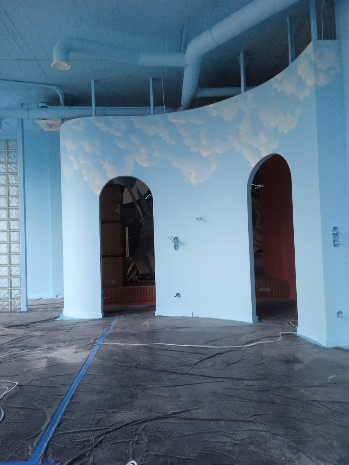 Niebieskie ściany z malowanymi chmurami w biurze, podłoga zabezpieczona folią malarską, widoczne instalacje pod sufitem.