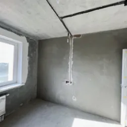 Surowy stan wykończenia wnętrza: widoczne przewody elektryczne na suficie, przygotowane punkty instalacyjne na ścianie, okno z grzejnikiem i ślady po gładzi na ścianach.