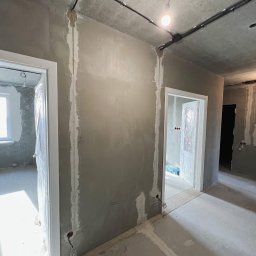 ELEKTRIKVATER SPÓŁKA Z OGRANICZONĄ ODPOWIEDZIALNOŚCIĄ - Surowe wnętrze w trakcie prac remontowych: ściany pokryte szarym tynkiem, widoczne bruzdy pod instalacje elektryczne, prowizoryczne oświetlenie żarówką, drzwi zabezpieczone folią. W tle kolejne...