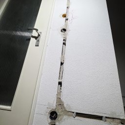 ELEKTRIKVATER SPÓŁKA Z OGRANICZONĄ ODPOWIEDZIALNOŚCIĄ - Biała ściana z wyżłobionym pionowym kanałem, w którym umieszczono peszle z przewodami elektrycznymi; widoczne otwory pod gniazdka i puszki elektryczne, ściana po bruzdowaniu wymaga wykończenia.