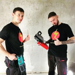 ELEKTRIKVATER SPÓŁKA Z OGRANICZONĄ ODPOWIEDZIALNOŚCIĄ - Dwóch elektryków w koszulkach z logiem firmy, jeden trzyma próbnik napięcia i wiertarkę, drugi bruzdownicę, na tle surowej ściany.