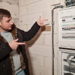 ELEKTRIKVATER SPÓŁKA Z OGRANICZONĄ ODPOWIEDZIALNOŚCIĄ - Mężczyzna w kurtce zimowej wskazuje na białą rozdzielnicę elektryczną ABB na ścianie z betonu komórkowego w surowym stanie.