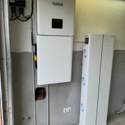 Zestaw akumulatorów energii z falownikiem marki QCells zamontowany na surowej ścianie z widocznymi przewodami i gniazdami elektrycznymi.