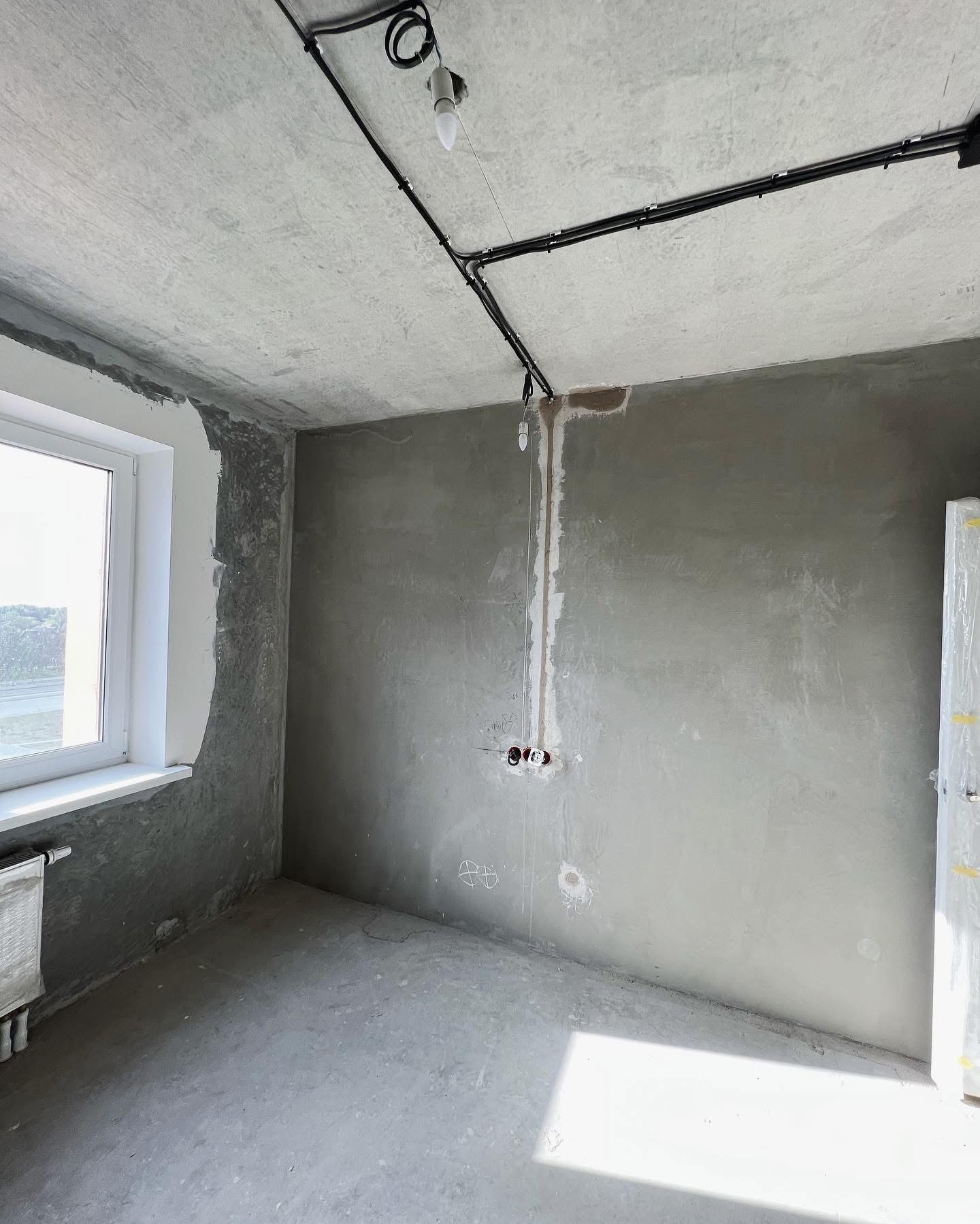 Surowy stan wykończenia wnętrza: widoczne przewody elektryczne na suficie, przygotowane punkty instalacyjne na ścianie, okno z grzejnikiem i ślady po gładzi na ścianach.