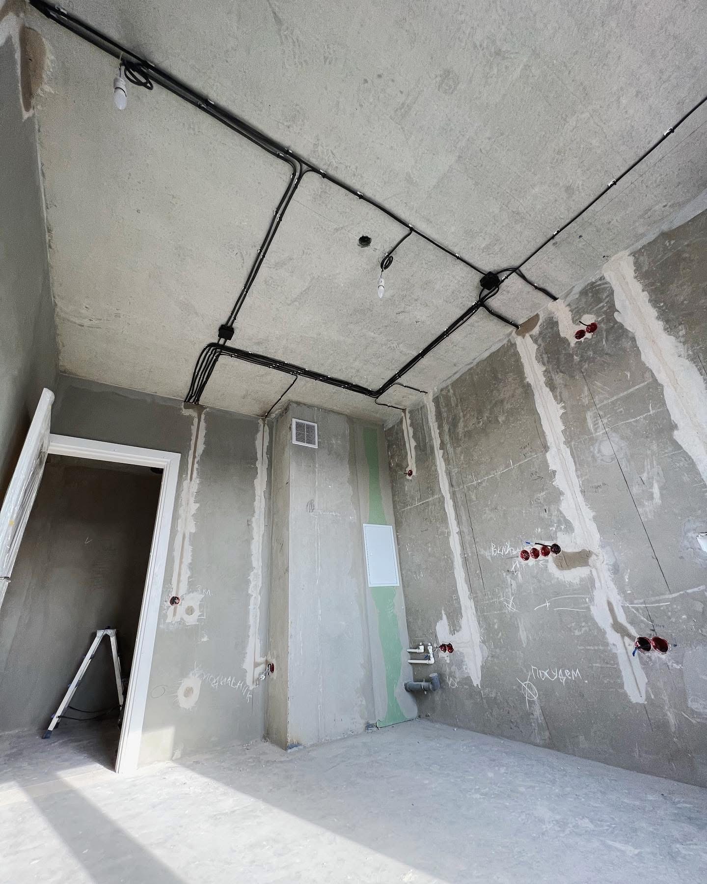 Surowy pokój w trakcie remontu z widocznymi przewodami elektrycznymi na suficie, puszkami instalacyjnymi w ścianach, częściowo zagruntowanymi ścianami i drabiną opartą o futrynę drzwiową.