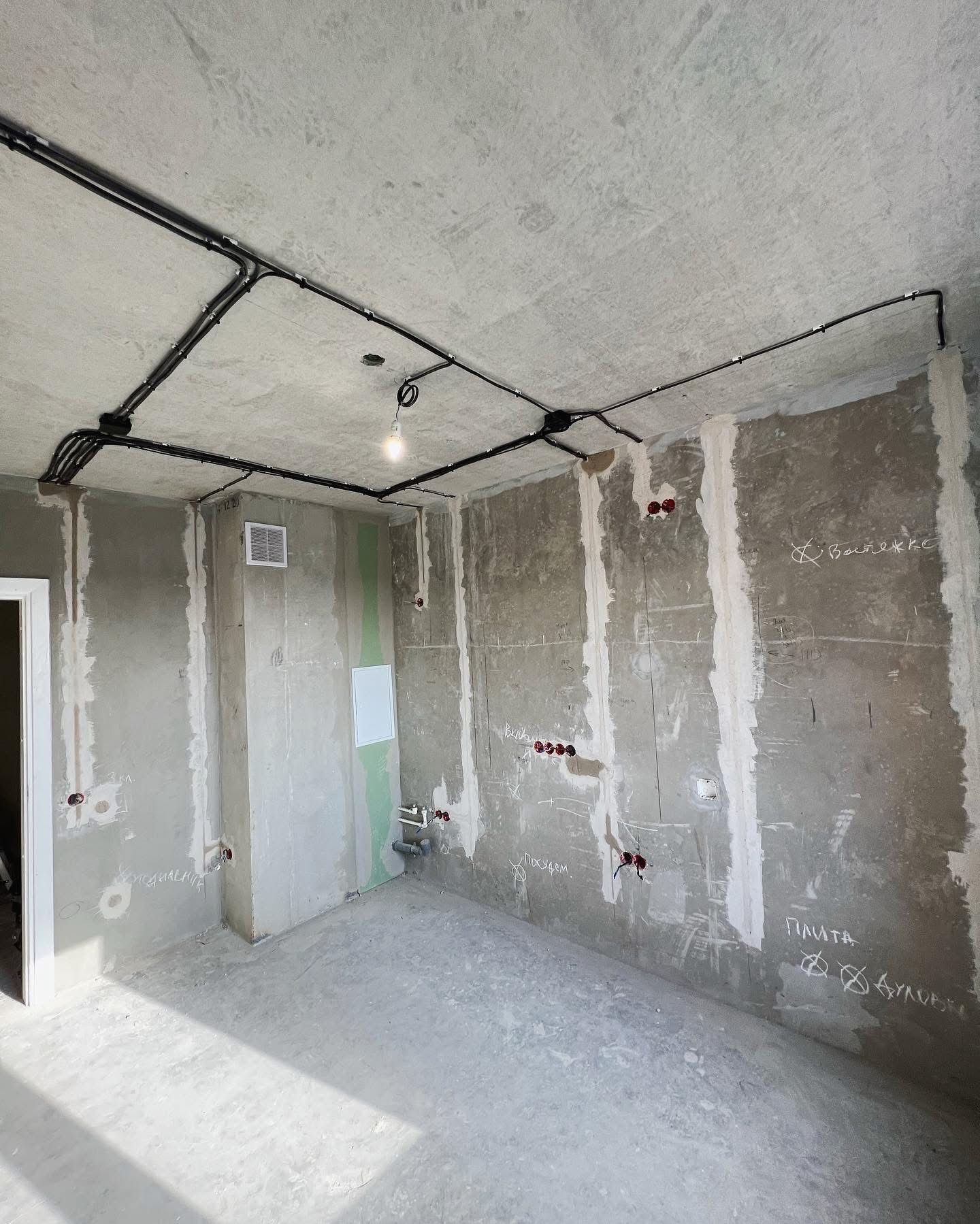 Surowe wnętrze w stanie deweloperskim z widoczną instalacją elektryczną: czarne kable prowadzone po suficie i ścianach, gniazdka w puszkach, surowy beton, ślady po tynku, pojedyncza żarówka na kablu.
