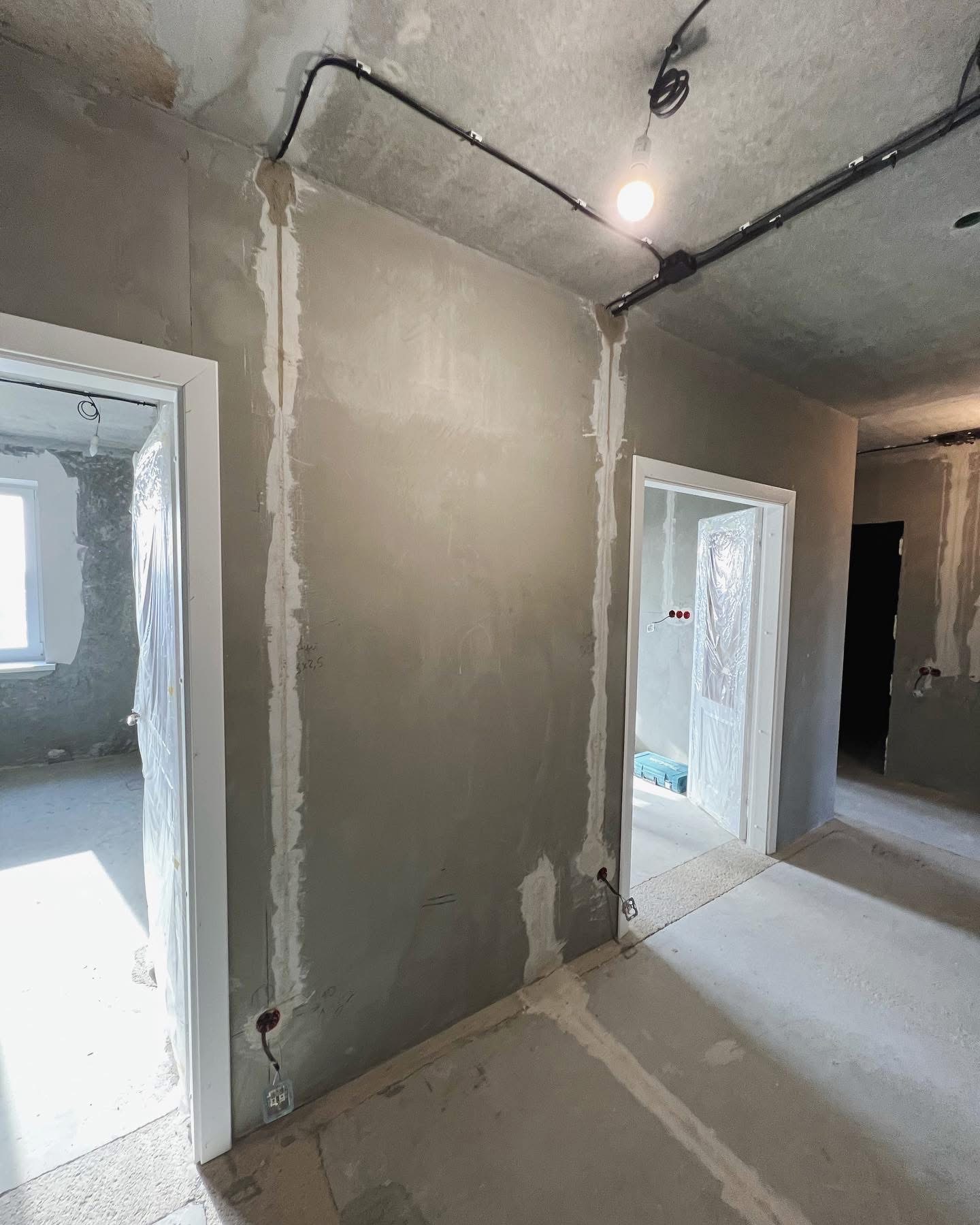 Surowe wnętrze w trakcie prac remontowych: ściany pokryte szarym tynkiem, widoczne bruzdy pod instalacje elektryczne, prowizoryczne oświetlenie żarówką, drzwi zabezpieczone folią. W tle kolejne...