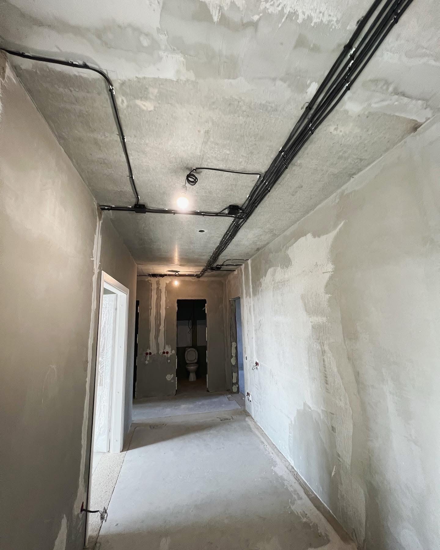 Wnętrze w stanie deweloperskim z widocznymi przewodami elektrycznymi poprowadzonymi po suficie, surowe ściany i podłoga, w tle widoczna toaleta.