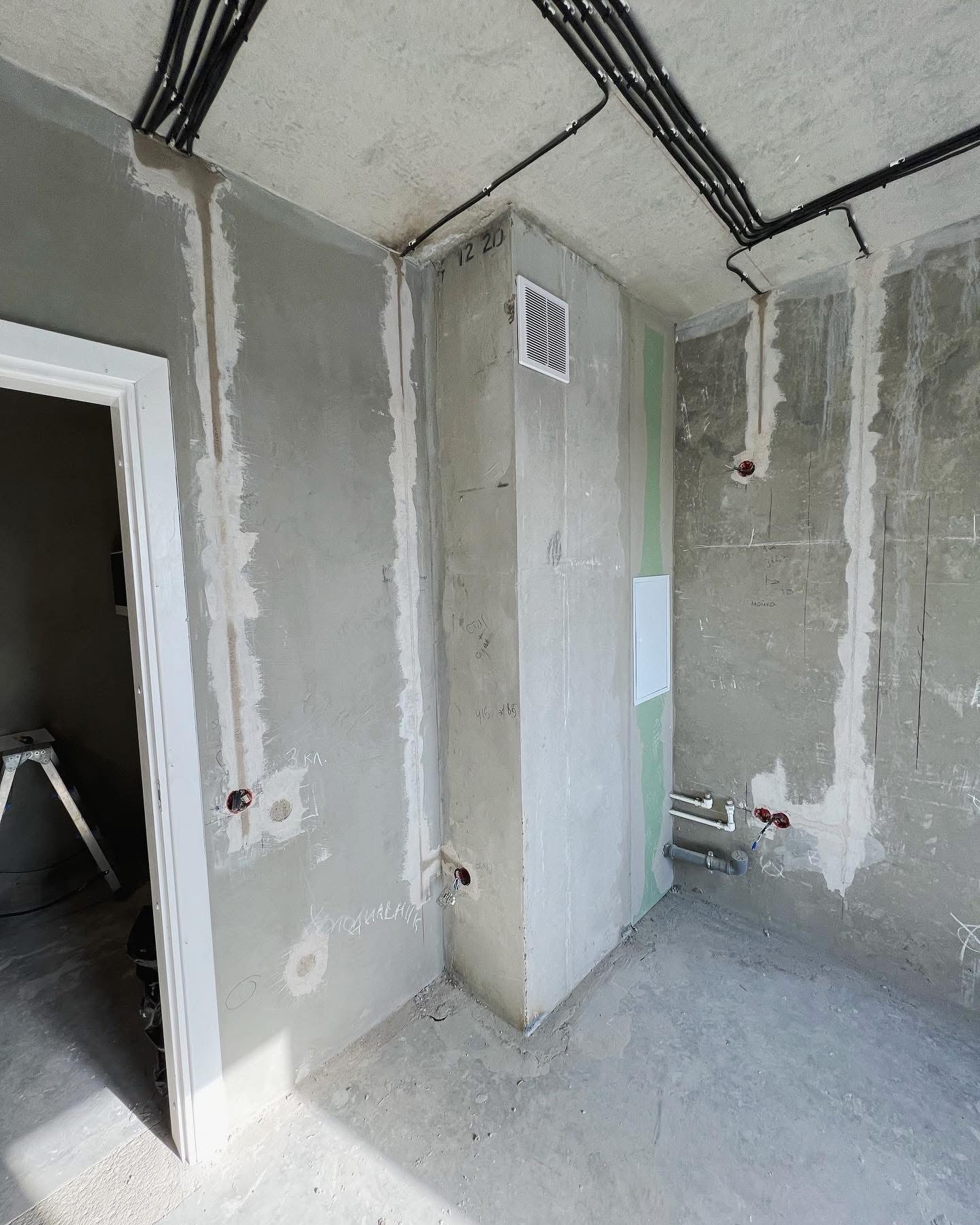 Surowe wnętrze w trakcie remontu: ściany z szarym tynkiem, widoczne instalacje elektryczne i rury, czarne kable pod sufitem, biała kratka wentylacyjna i ościeżnica drzwiowa, podłoga wylewka...