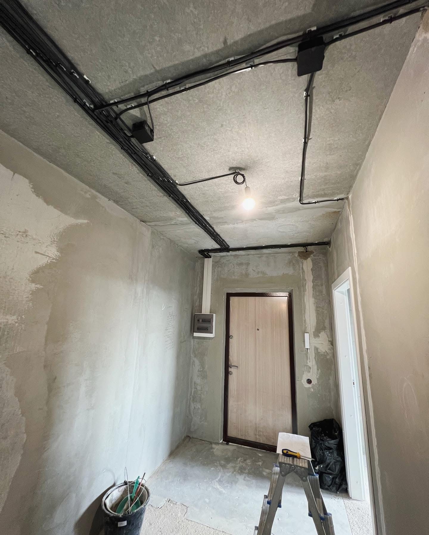 Wnętrze w trakcie remontu: surowy beton, poprowadzone czarne kable elektryczne pod sufitem, wisząca żarówka, drabina z narzędziami, ściany przygotowane do malowania.