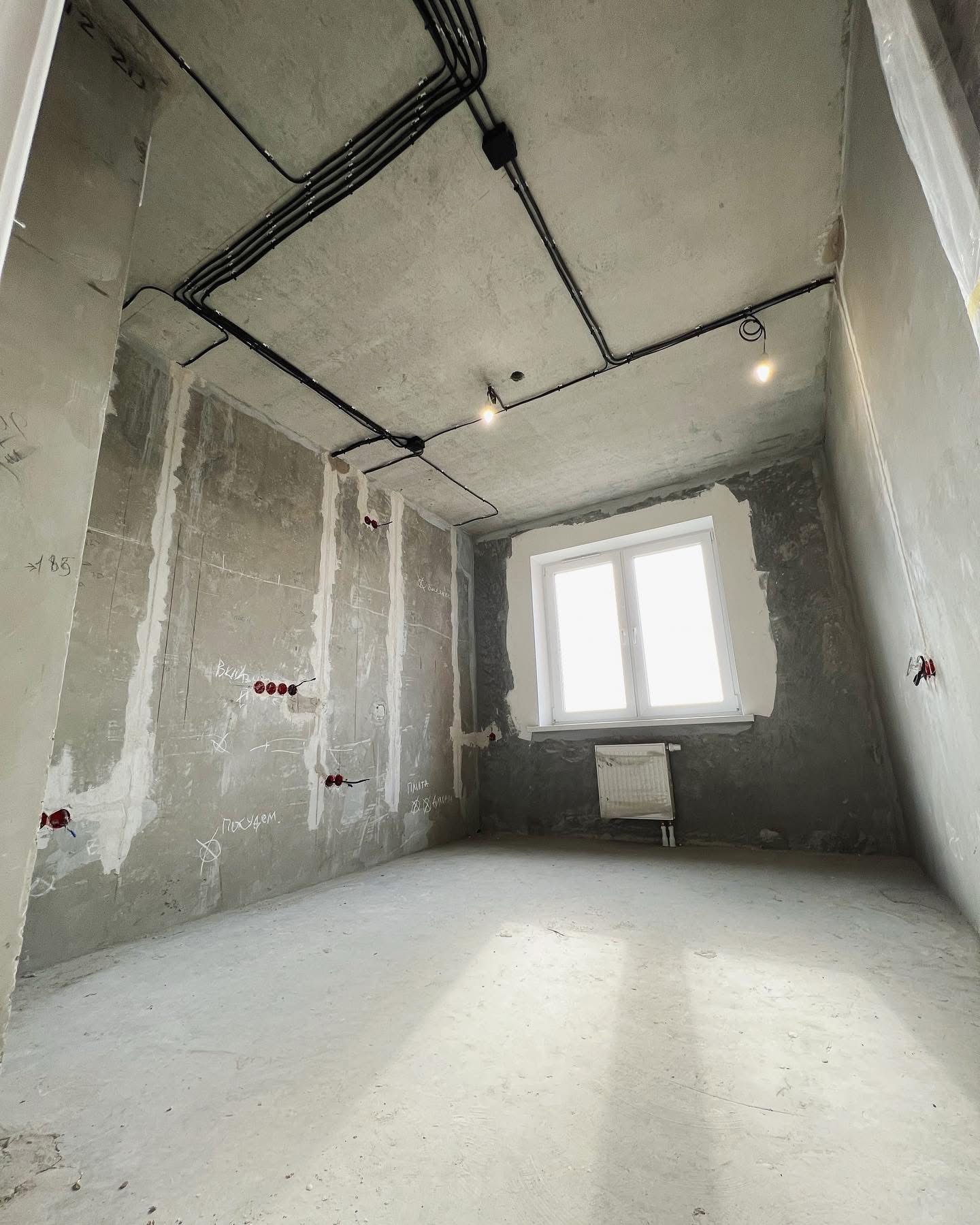 Surowe wnętrze w trakcie remontu: widoczne kable elektryczne pod sufitem, instalacja elektryczna w ścianach, surowe ściany z notatkami i białe okno z grzejnikiem.