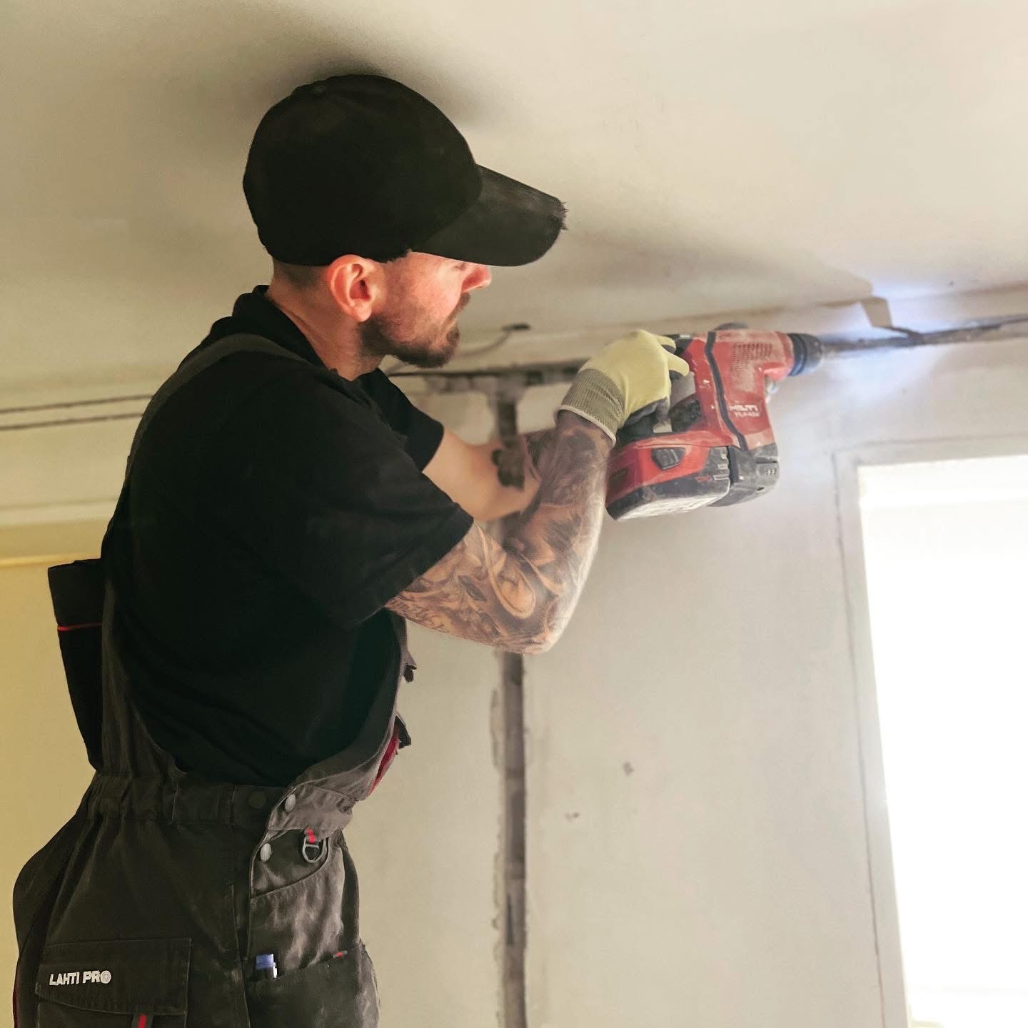 Mężczyzna z tatuażem na ramieniu, w czarnej czapce i kombinezonie Lahti Pro, wierci czerwoną wiertarką Hilti w suficie w trakcie prac remontowych.