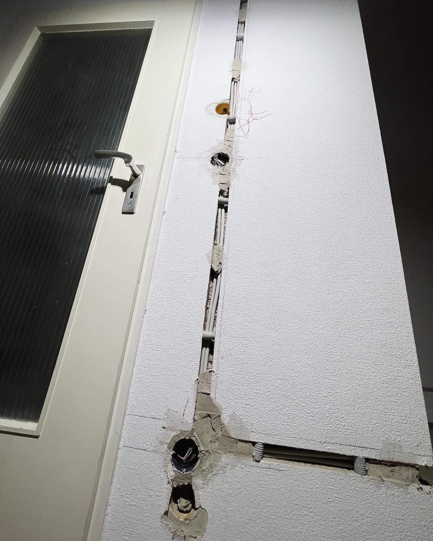 Biała ściana z wyżłobionym pionowym kanałem, w którym umieszczono peszle z przewodami elektrycznymi; widoczne otwory pod gniazdka i puszki elektryczne, ściana po bruzdowaniu wymaga wykończenia.