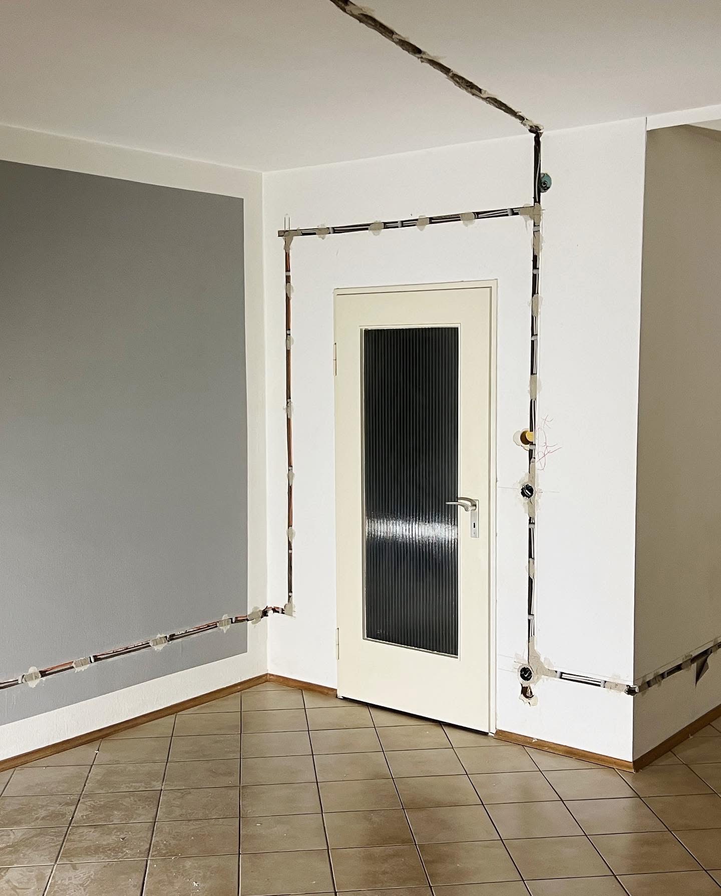 Przygotowanie instalacji elektrycznej w pomieszczeniu: widoczne kable przymocowane do ściany za pomocą uchwytów, bruzdy w ścianie i suficie, oraz otwory pod gniazdka elektryczne obok białych drzwi...