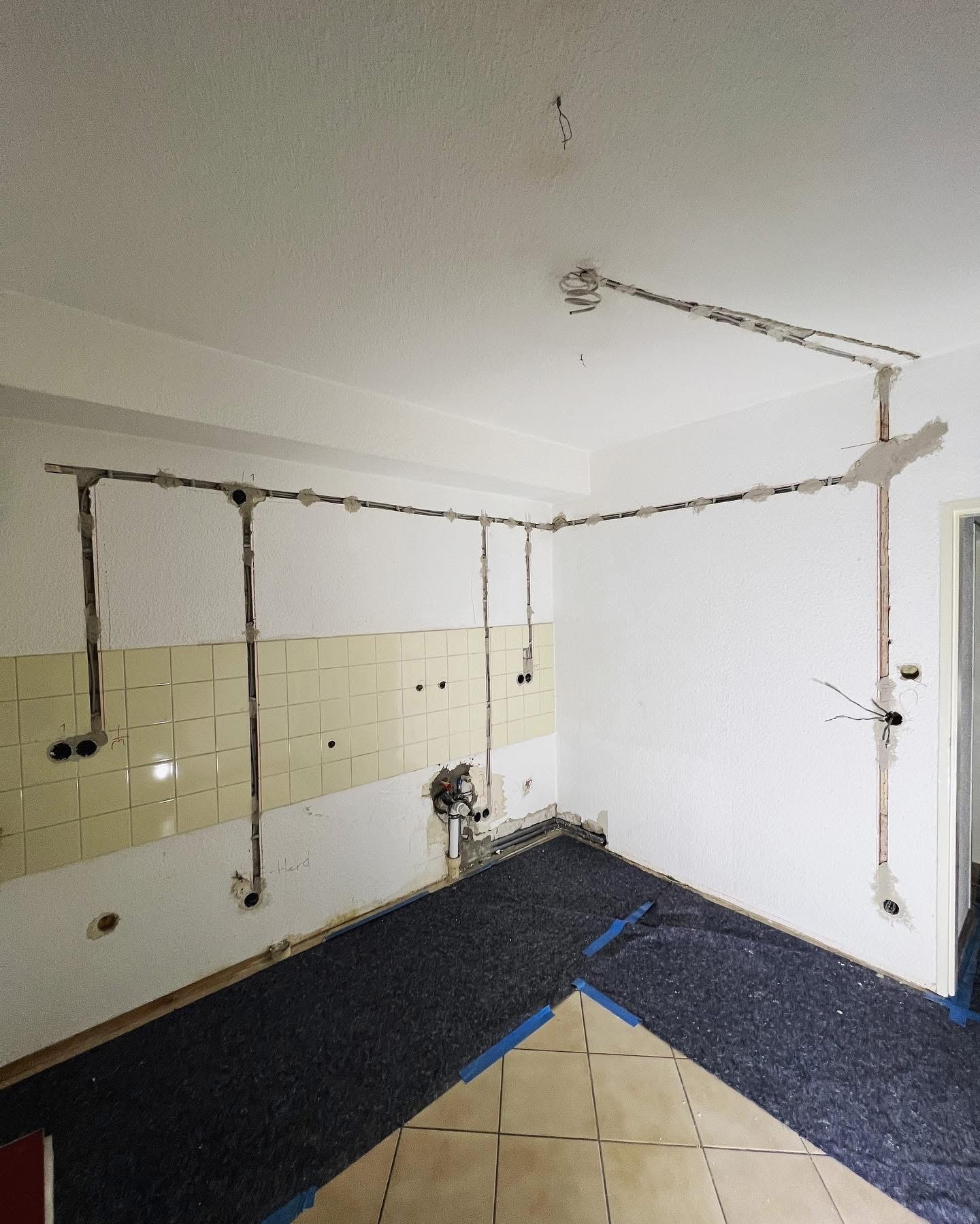 Kuchnia w trakcie remontu: widoczne nowe instalacje elektryczne prowadzone po ścianach i suficie, zabezpieczona podłoga, częściowo zdjęte płytki, przygotowania do prac wykończeniowych.