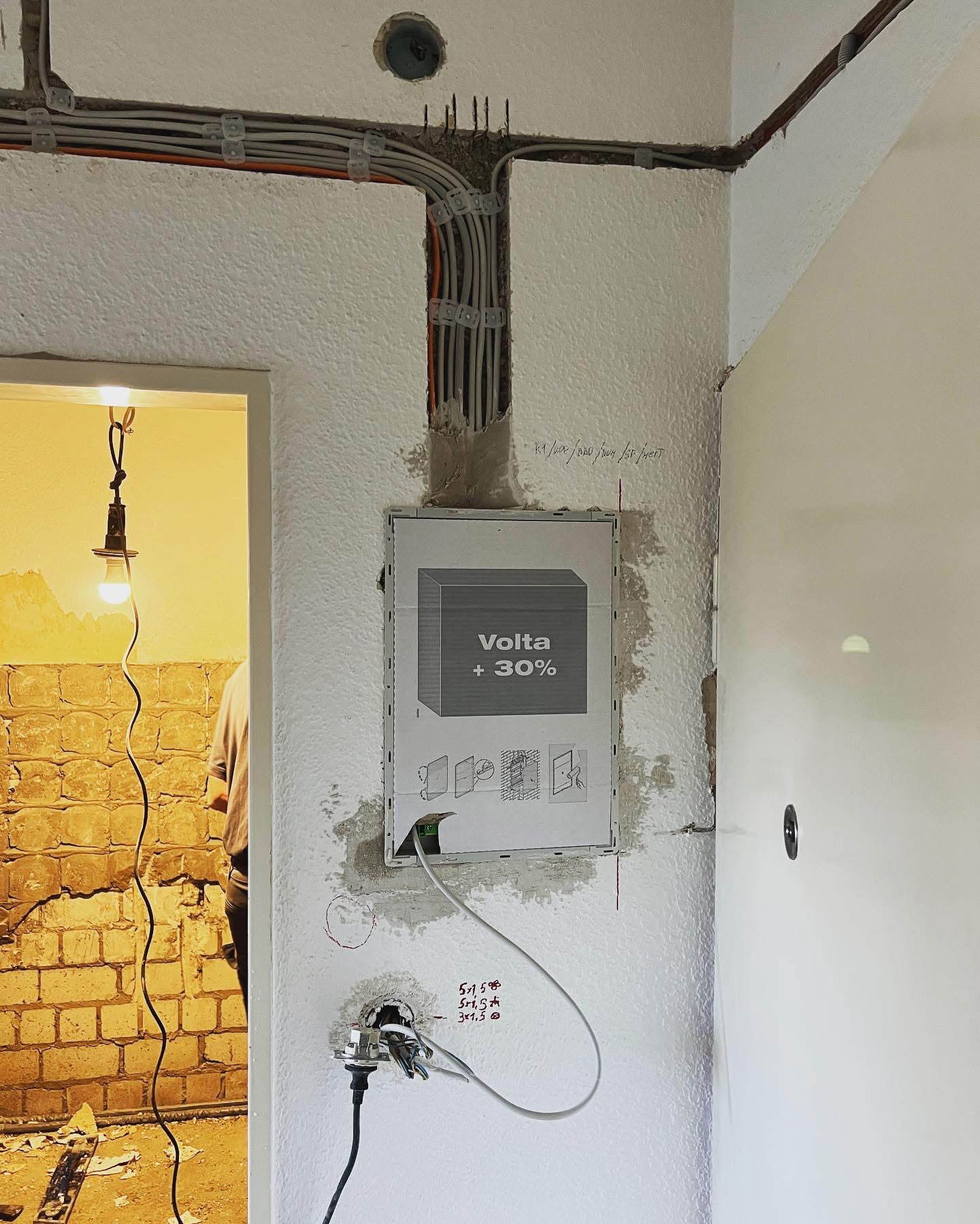 Instalacja elektryczna w trakcie montażu, widoczne kable prowadzone w ścianie, skrzynka rozdzielcza Volta +30% oraz gniazdko z wystającymi przewodami. W tle pomieszczenie w trakcie remontu...