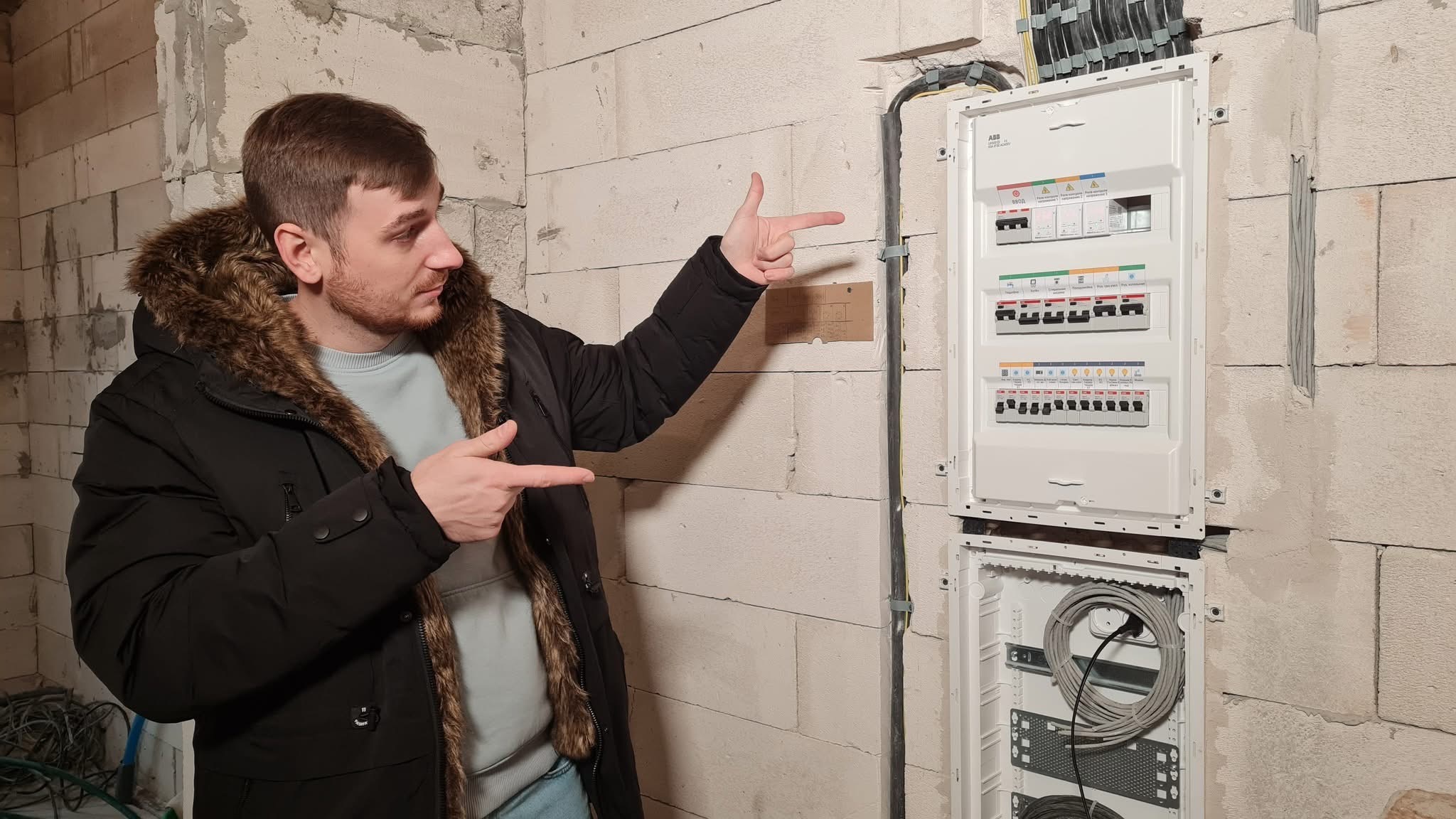 Mężczyzna w kurtce zimowej wskazuje na białą rozdzielnicę elektryczną ABB na ścianie z betonu komórkowego w surowym stanie.