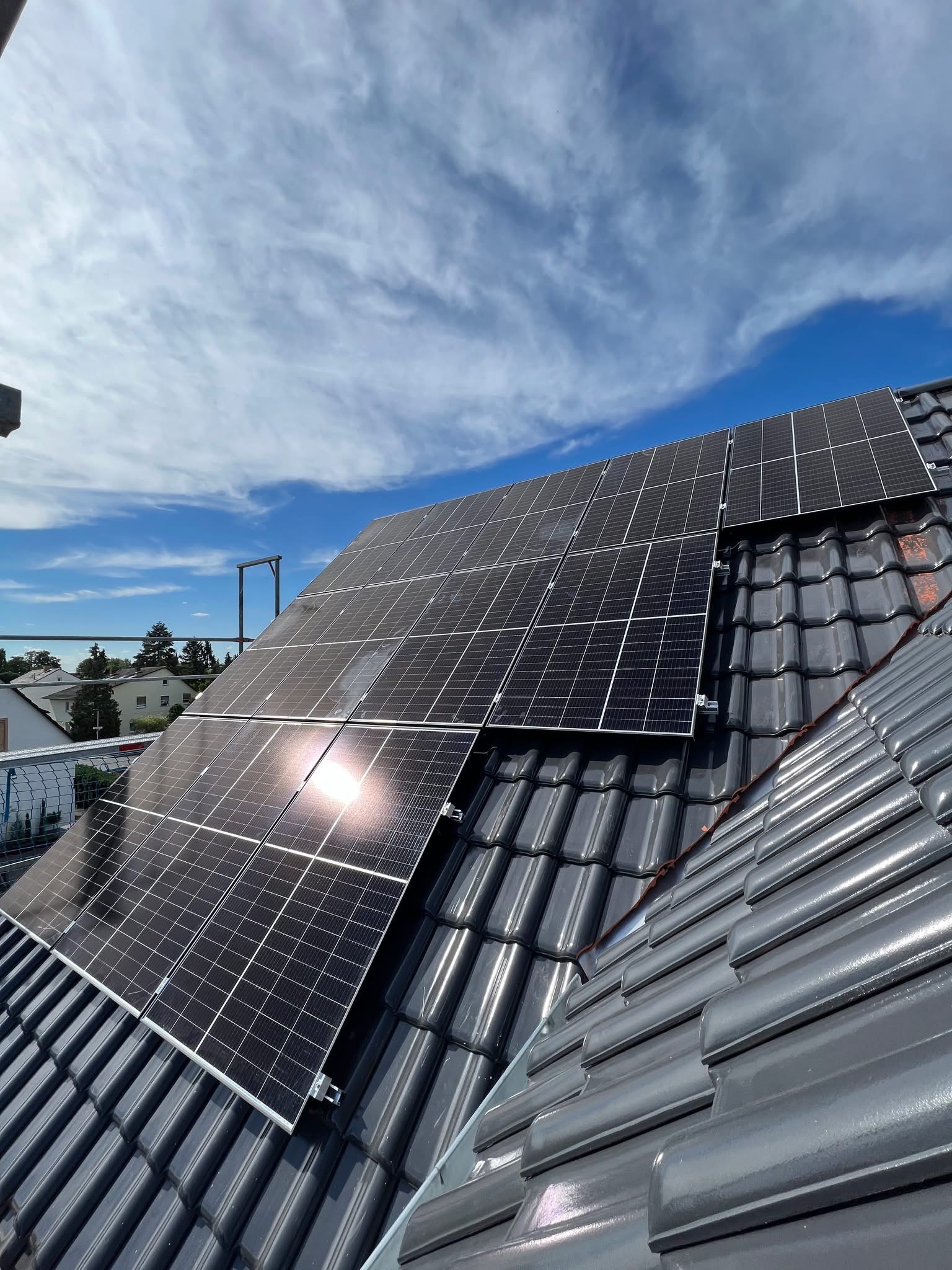 Panele słoneczne zamontowane na ciemnoszarym dachu z dachówki ceramicznej, odbijające słońce na tle błękitnego nieba z chmurami.