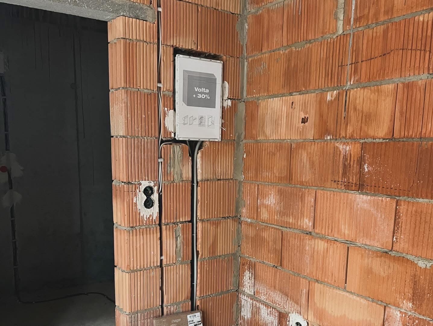 Instalacja elektryczna w trakcie montażu: przewody prowadzone w bruzdach w ścianie z cegły, puszka elektryczna widoczna po prawej stronie, tablica rozdzielcza z napisem 'Volta +30%' zamontowana...