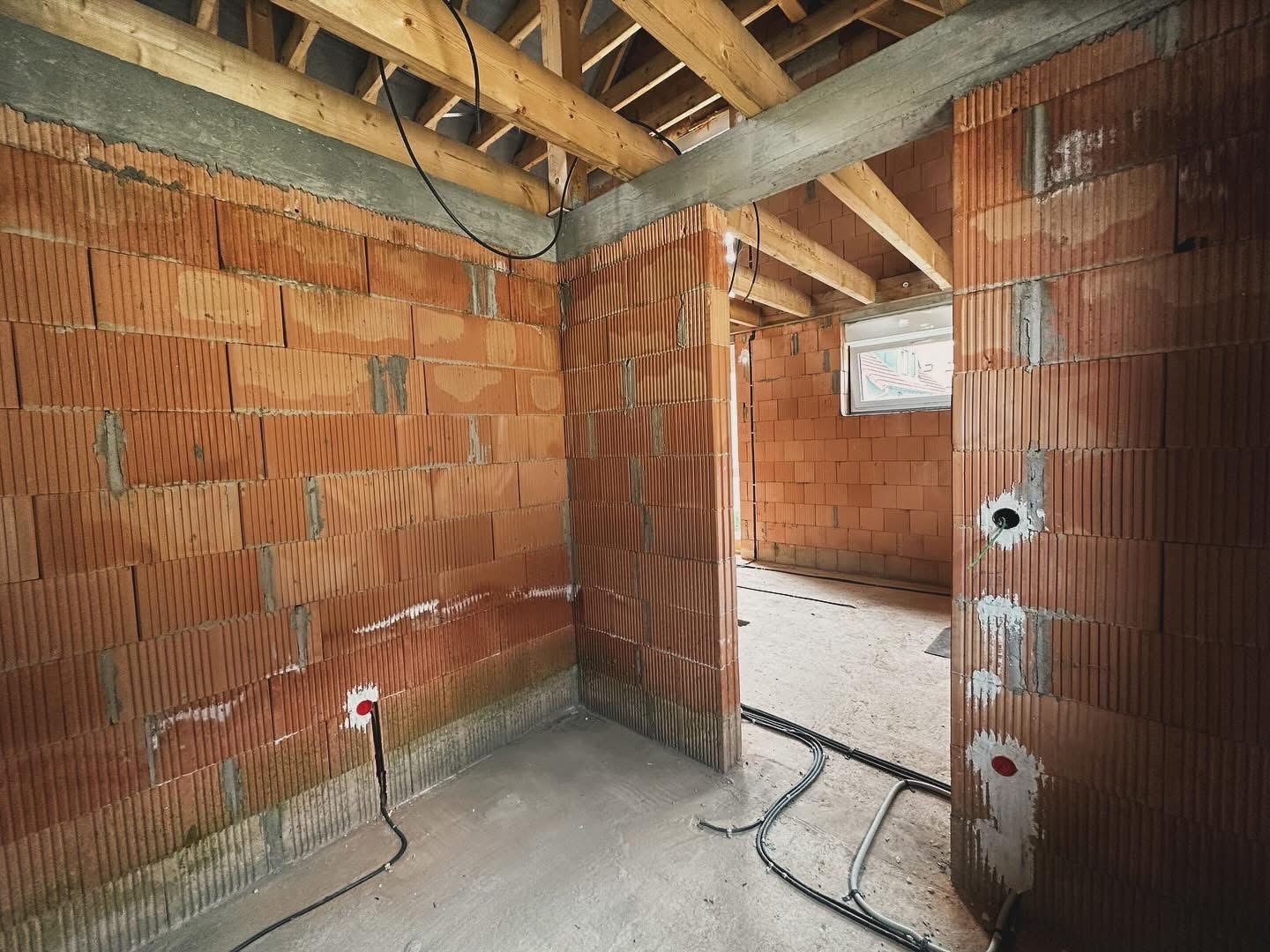 Wnętrze w trakcie budowy: surowe ściany z czerwonej cegły, drewniana konstrukcja dachu, w ścianach widoczne otwory na instalacje elektryczne, kable leżące na surowej podłodze.