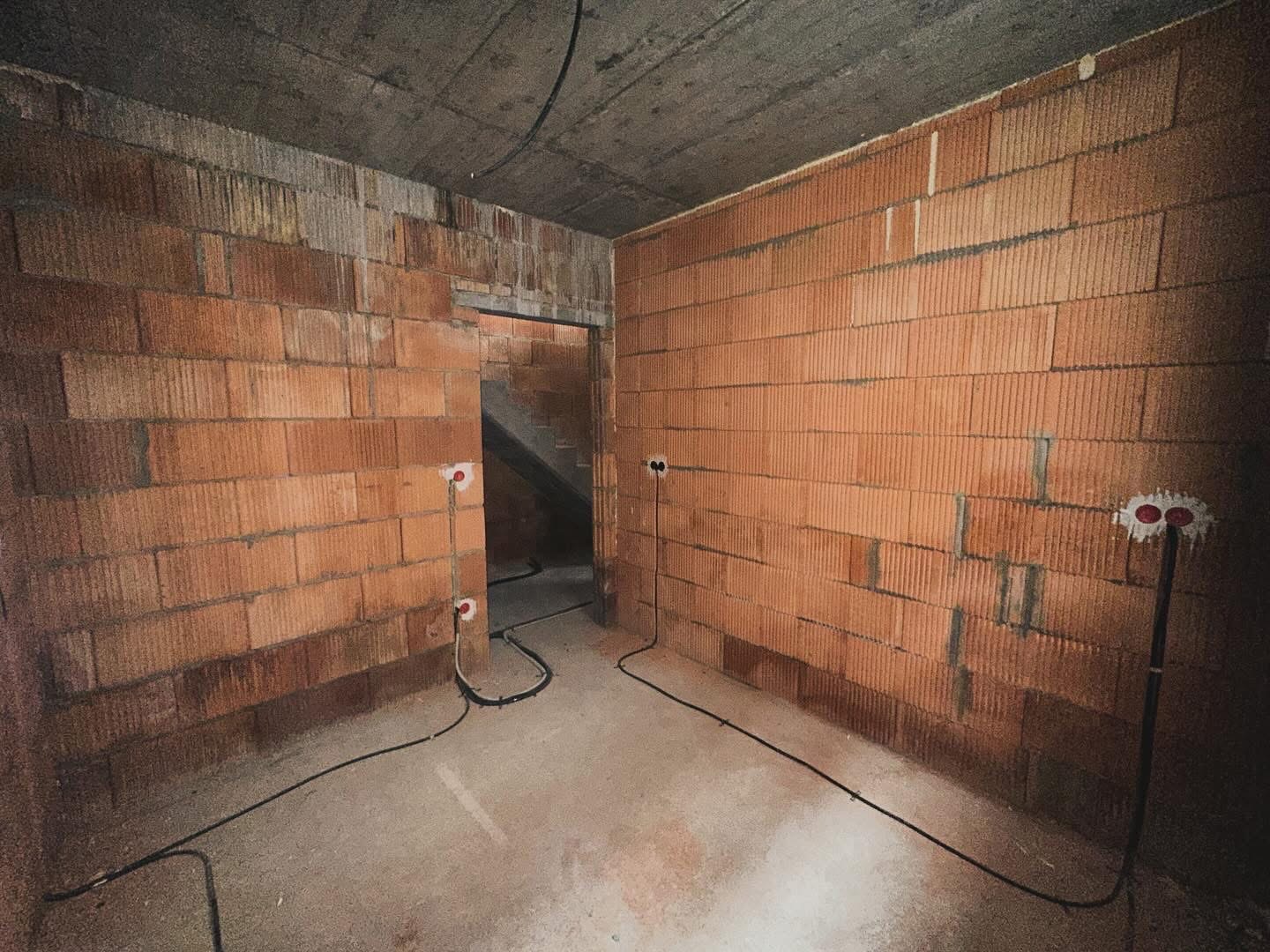 Surowy stan wnętrza w Warszawie: ściany z czerwonej cegły, betonowy sufit, wystające kable elektryczne z puszkami instalacyjnymi, widok na schody w głębi.