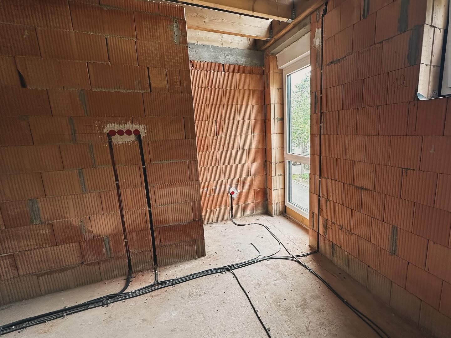 Wnętrze w stanie surowym z widocznymi przewodami elektrycznymi w ścianie z czerwonej cegły dziurawki oraz kablami prowadzonymi po podłodze, okno w tle.