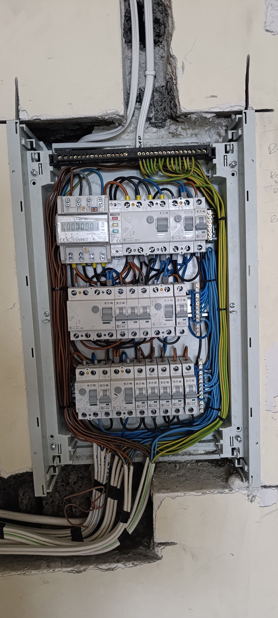 Rozdzielnica elektryczna w trakcie montażu, widoczne zabezpieczenia, przewody i licznik energii, osadzona w ścianie z widocznymi śladami prac budowlanych.