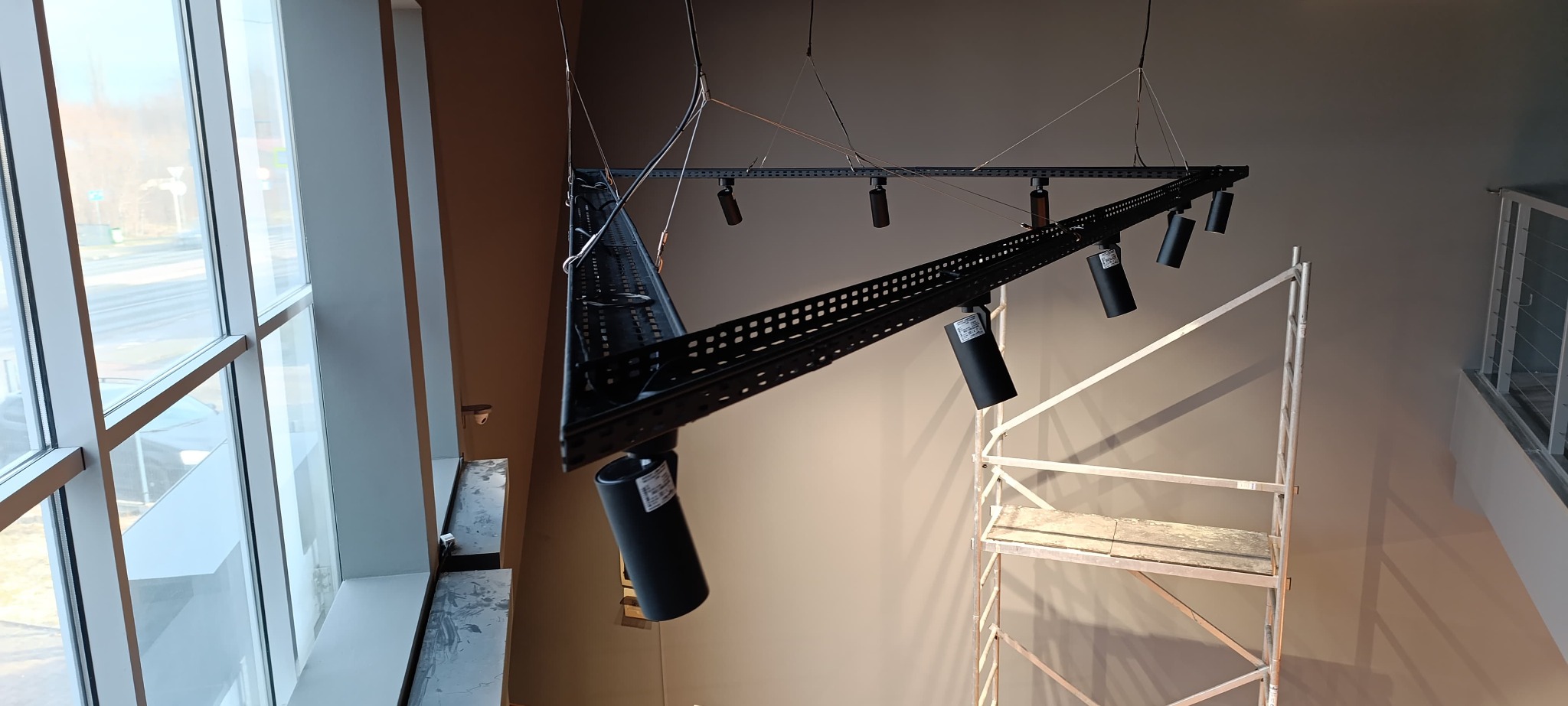 Montaż czarnej, metalowej konstrukcji oświetleniowej z reflektorami w wysokim pomieszczeniu, widoczne rusztowanie i duże okno.