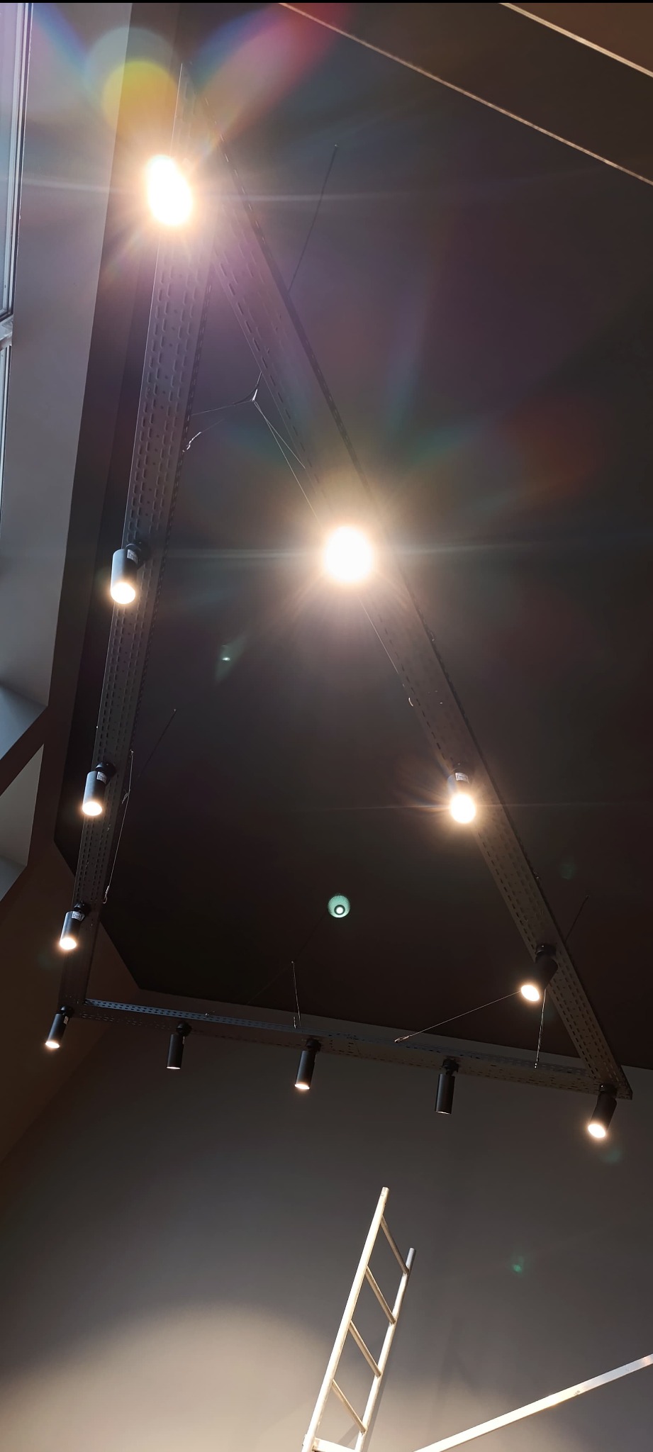 Oświetlenie sufitowe z reflektorami na szynoprzewodach w Tychach, widoczne z perspektywy podłogi, z fragmentem białej drabiny malarskiej w dolnym rogu.