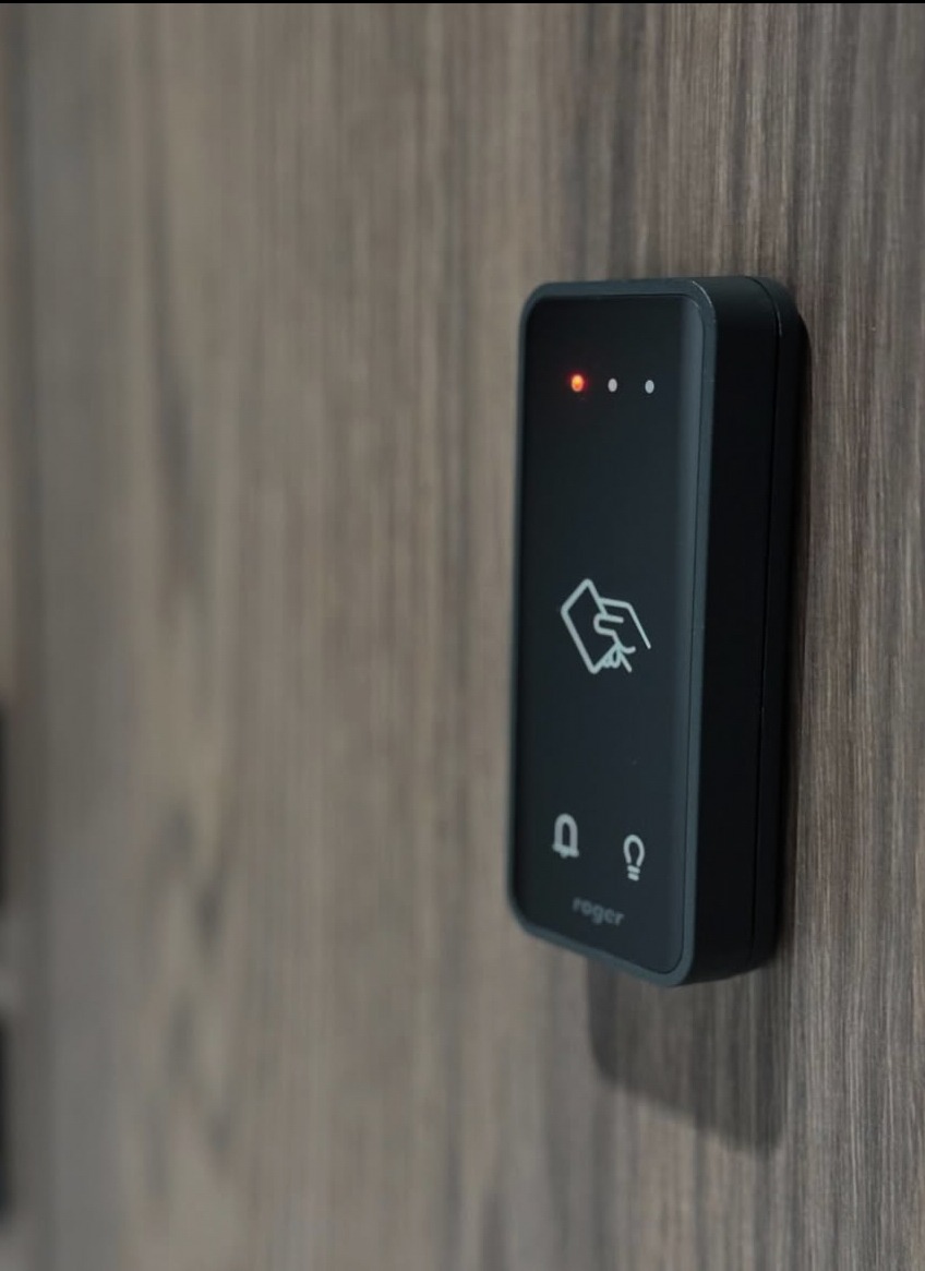 Czarny panel kontrolny systemu dostępu Roger zamontowany na drewnianej ścianie, z ikonami karty, dzwonka i żarówki, oraz trzema diodami LED.