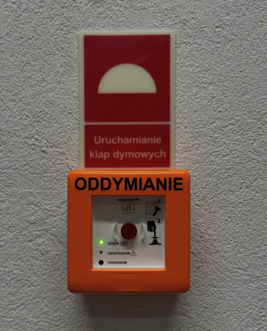 Przycisk oddymiania w pomarańczowej obudowie z instrukcją, zamontowany na szarej ścianie obok znaku informującego o uruchamianiu klap dymowych.