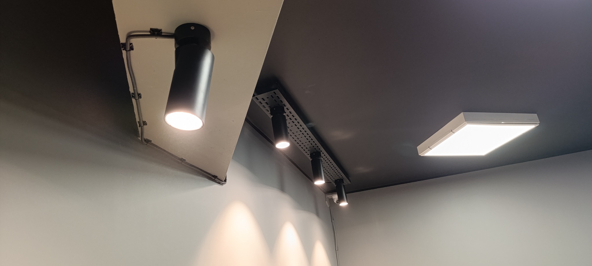 Zamontowane czarne reflektory punktowe na suficie z szynoprzewodem i natynkowa lampa LED, widoczne kable i mocowania instalacji elektrycznej.