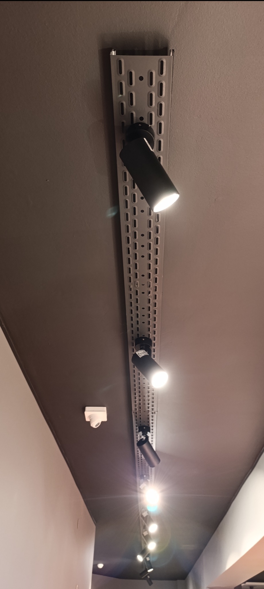 Szyna oświetleniowa z reflektorami punktowymi, zamontowana na suficie w szarym odcieniu, widoczne także elementy instalacji alarmowej.