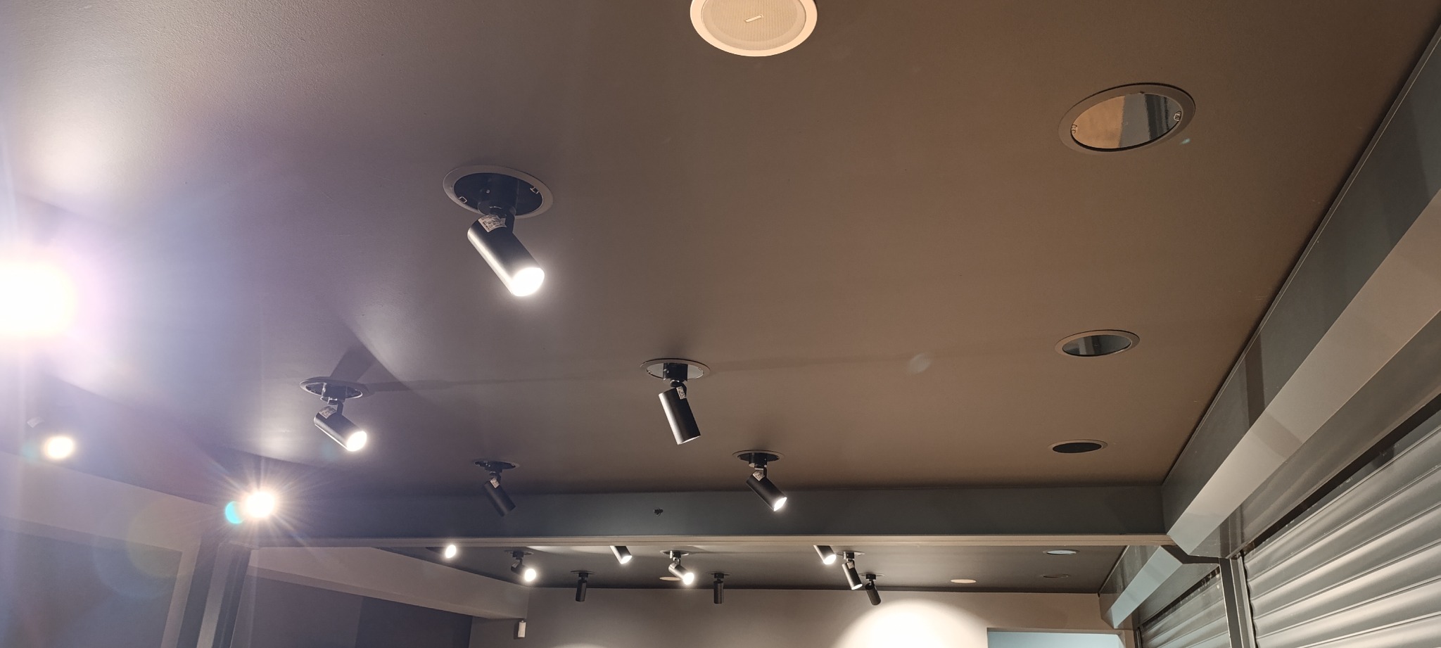 Sufit z zamontowanymi reflektorami punktowymi i głośnikami sufitowymi, widoczne także otwory instalacyjne, oświetlenie skierowane w dół.