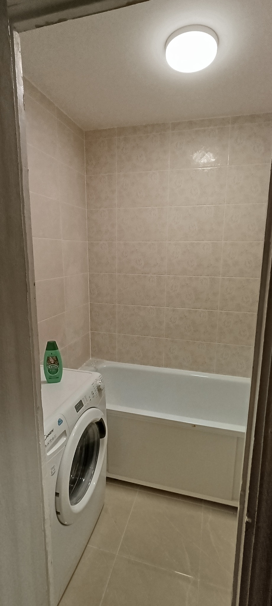 Jasna łazienka z beżowymi kafelkami, białą wanną i pralką Candy z zielonym szamponem na górze, oświetlona okrągłą lampą sufitową.