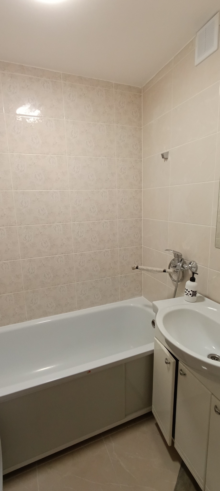 Wykończona łazienka z beżowymi, wzorzystymi płytkami, białą wanną i umywalką z szafką. Na ścianie widoczny uchwyt obłożony taśmą zabezpieczającą.