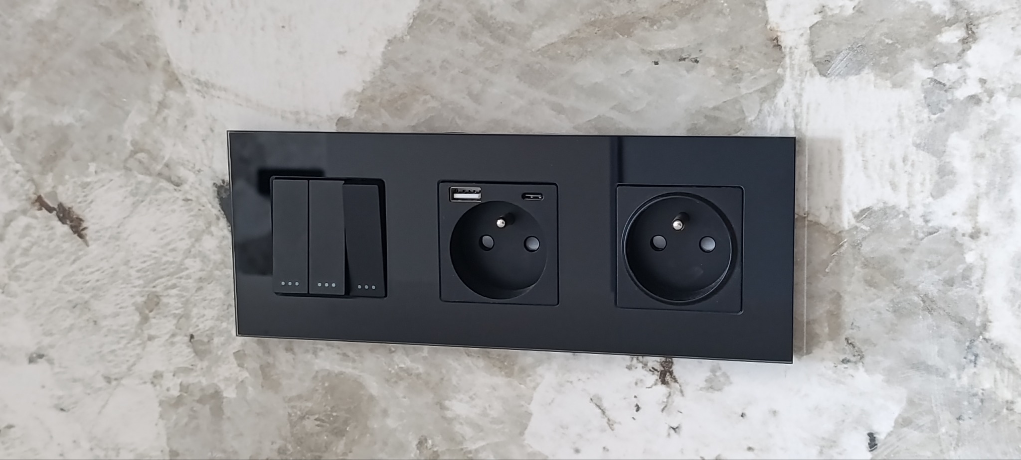 Czarny panel z trzema włącznikami i dwoma gniazdkami (jedno z USB) zamontowany na ścianie o marmurkowej fakturze. Nowoczesny design.