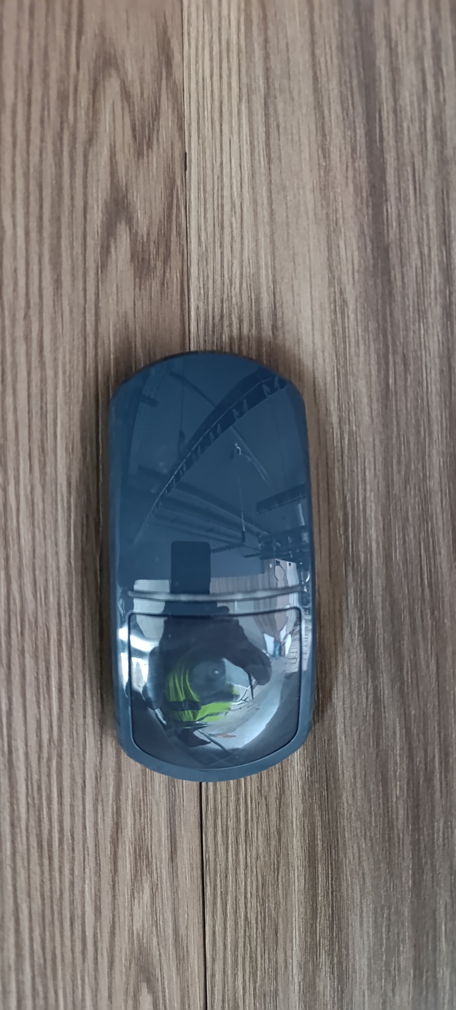 Zamontowany czujnik alarmowy w obudowie, widoczny na drewnianej powierzchni. W odbiciu czujnika widoczny fragment pomieszczenia i osoba w odblaskowej kamizelce.