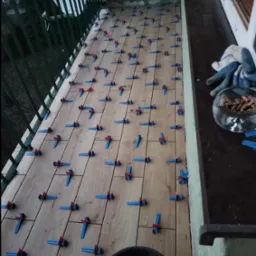 Układanie płytek drewnopodobnych na balkonie z użyciem niebiesko-czerwonych klipsów poziomujących, widoczny fragment wiadra i popielniczka z niedopałkami.