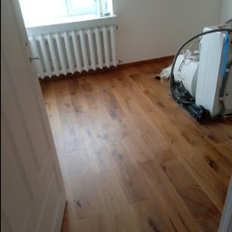Firma budowlana - Ukończona podłoga z paneli drewnianych w jasnym odcieniu, widoczny grzejnik pod oknem i sprzęt budowlany w rogu pokoju.