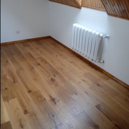 Firma budowlana - Jasne, drewniane panele podłogowe ułożone w jodełkę w pokoju na poddaszu z białymi ścianami, drewnianą boazerią na skosach dachu i białym grzejnikiem żeliwnym.