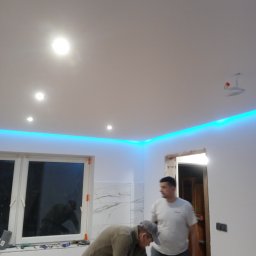 Firma budowlana - Prace wykończeniowe w pomieszczeniu: szlifowanie ściany przy oknie, widoczne oświetlenie LED podsufitowe, fragment marmurowej okładziny ściennej i mężczyzna w tle.