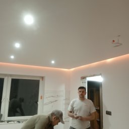 Firma budowlana - Dwaj mężczyźni w trakcie prac wykończeniowych w pomieszczeniu z oświetleniem LED podsufitowym i płytkami imitującymi marmur, jeden z nich pracuje przy oknie, drugi stoi w pobliżu otwartych drzwi.