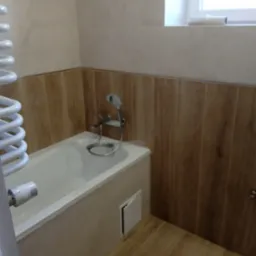 Wykończona łazienka z wanną obudowaną beżowymi płytkami, baterią wannową z prysznicem, grzejnikiem drabinkowym i panelami ściennymi imitującymi drewno.