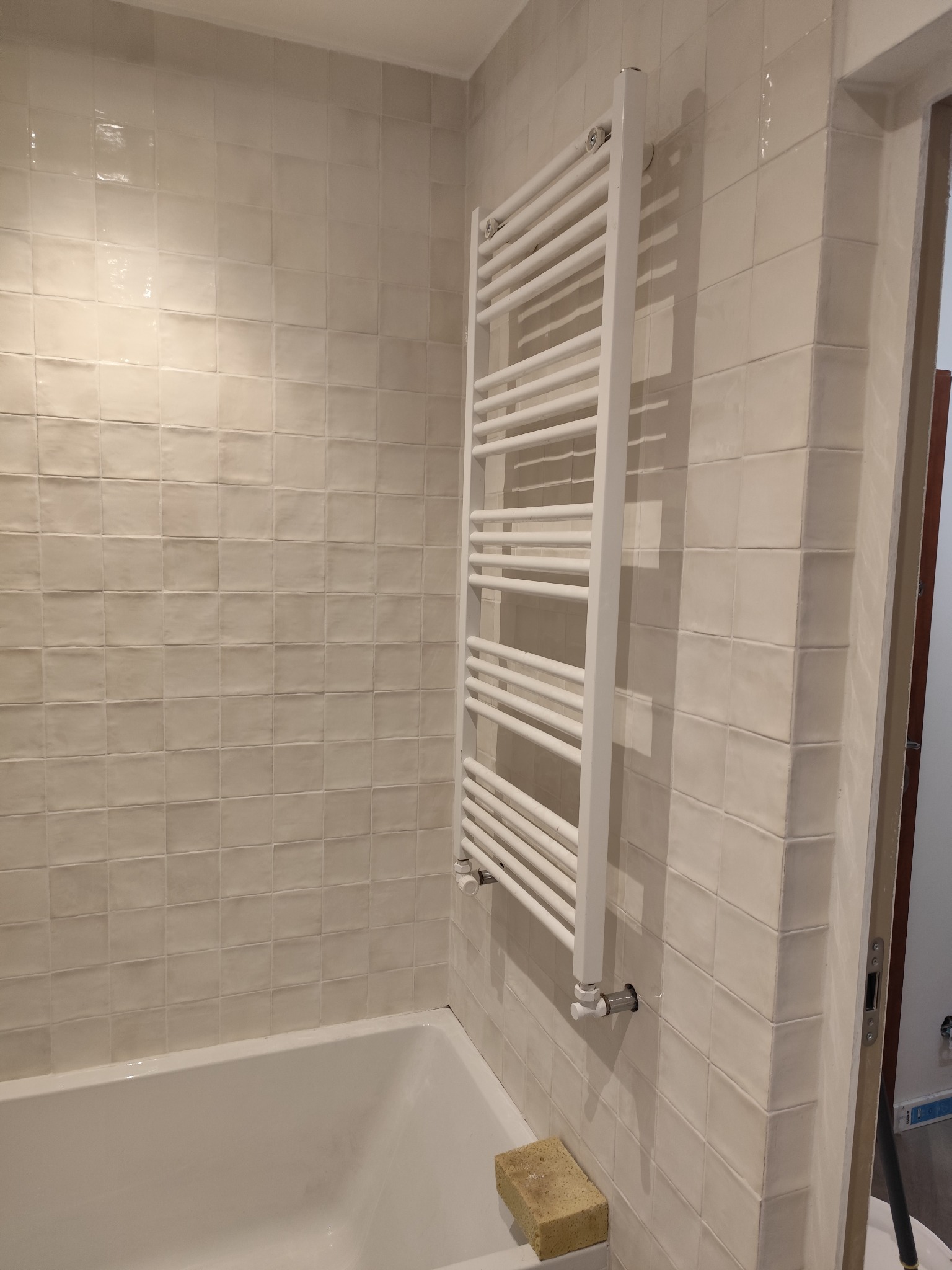 Biały grzejnik drabinkowy zamontowany na ścianie z kremowych, kwadratowych płytek w łazience, widoczna wanna i gąbka do czyszczenia.