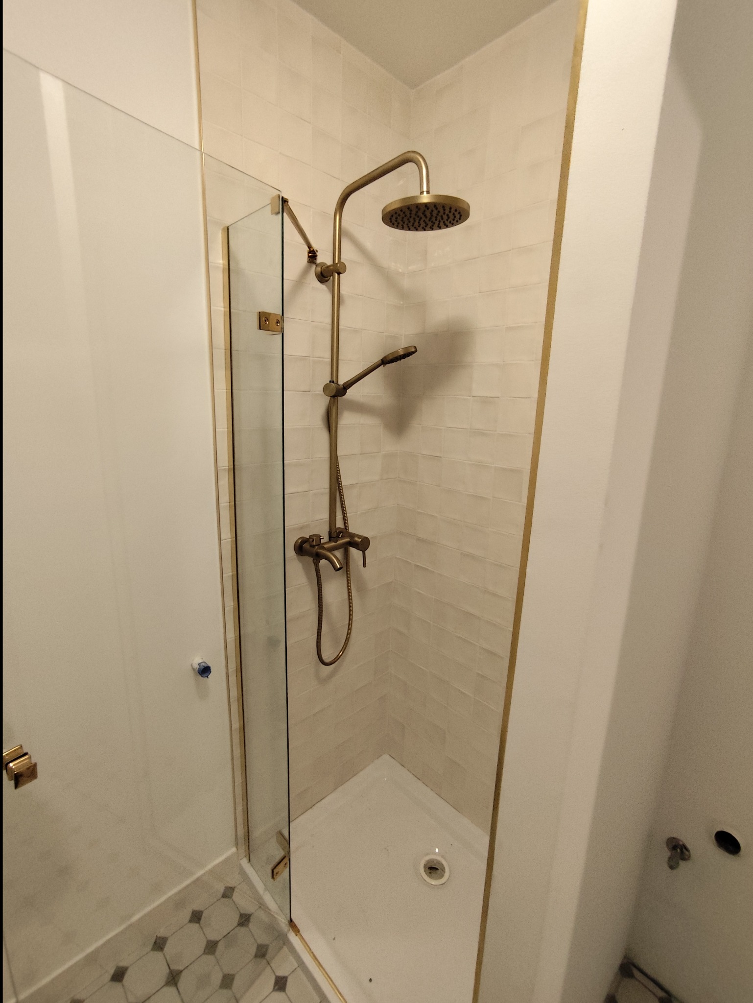 Wykończona kabina prysznicowa z armaturą w kolorze starego złota, beżowe płytki ułożone poziomo, szklane drzwi z złotymi ramami, podłoga wyłożona płytkami w geometryczny wzór.