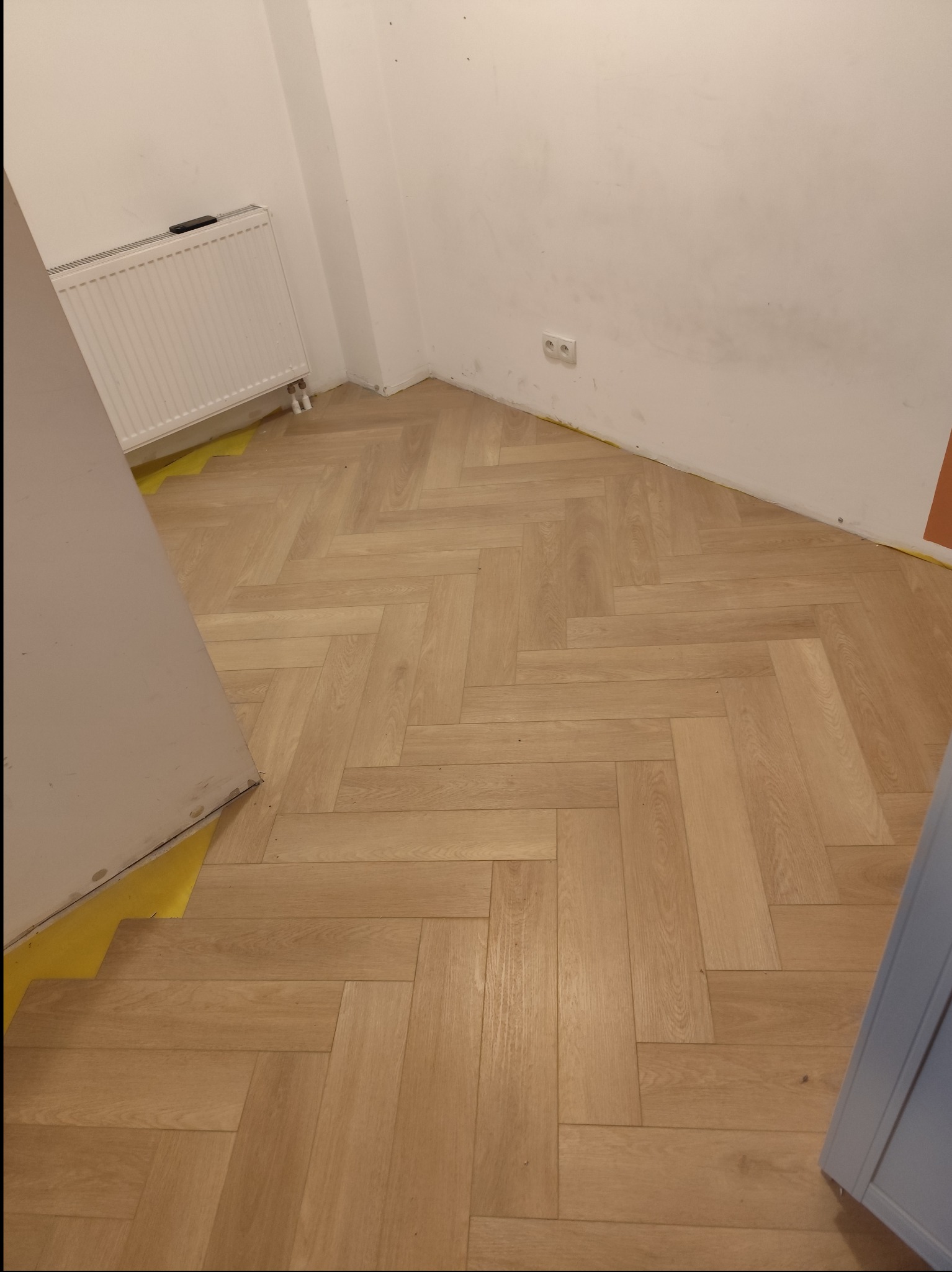 Ułożona parkietowa podłoga w jodełkę, zabezpieczona żółtą folią malarską przy ścianach i futrynie drzwi, widoczny grzejnik i gniazdko elektryczne.