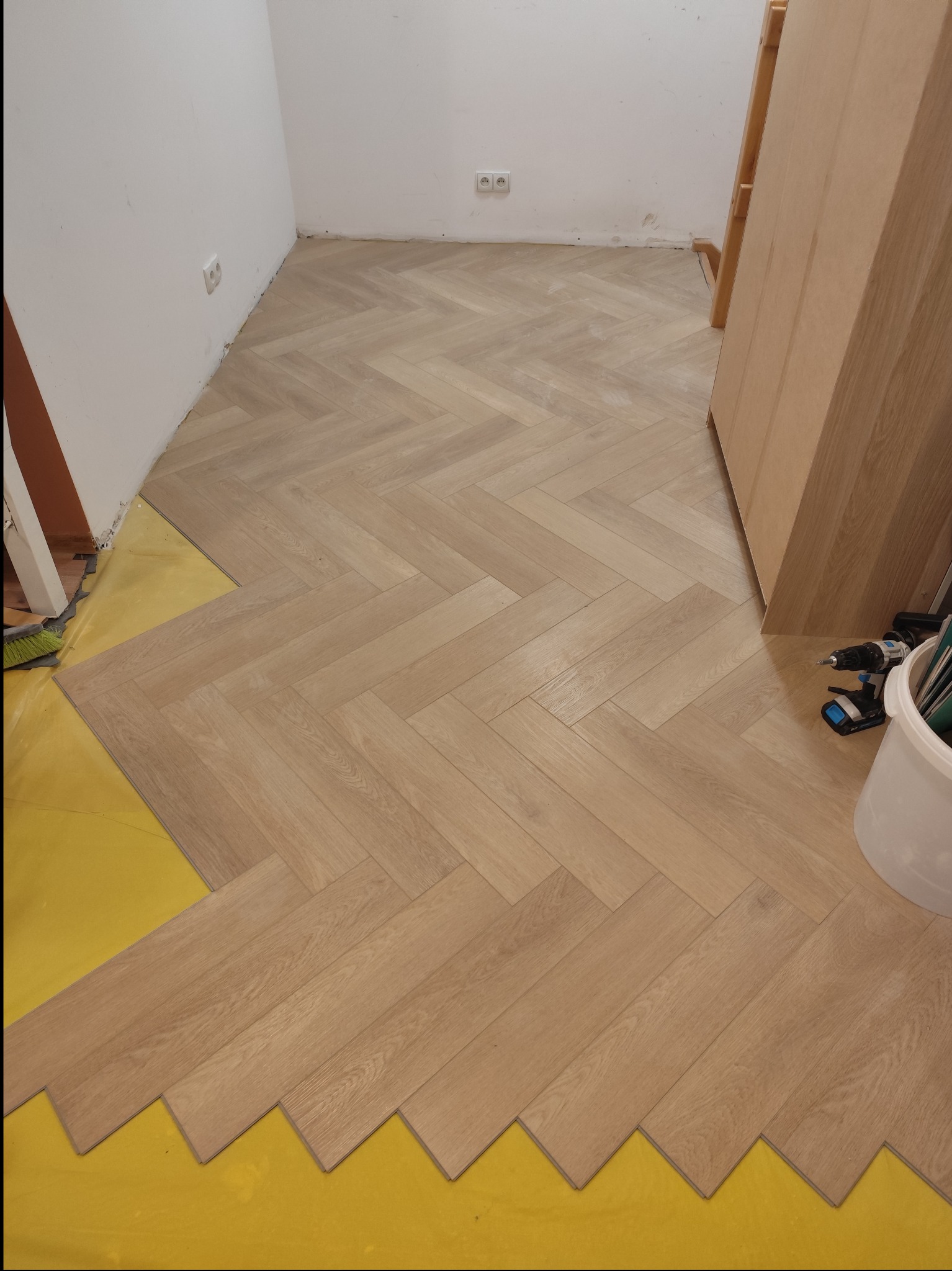 Układanie jasnej, drewnopodobnej podłogi w jodełkę na żółtym podkładzie, widoczne narzędzia pracy: wiertarka i wiadro.